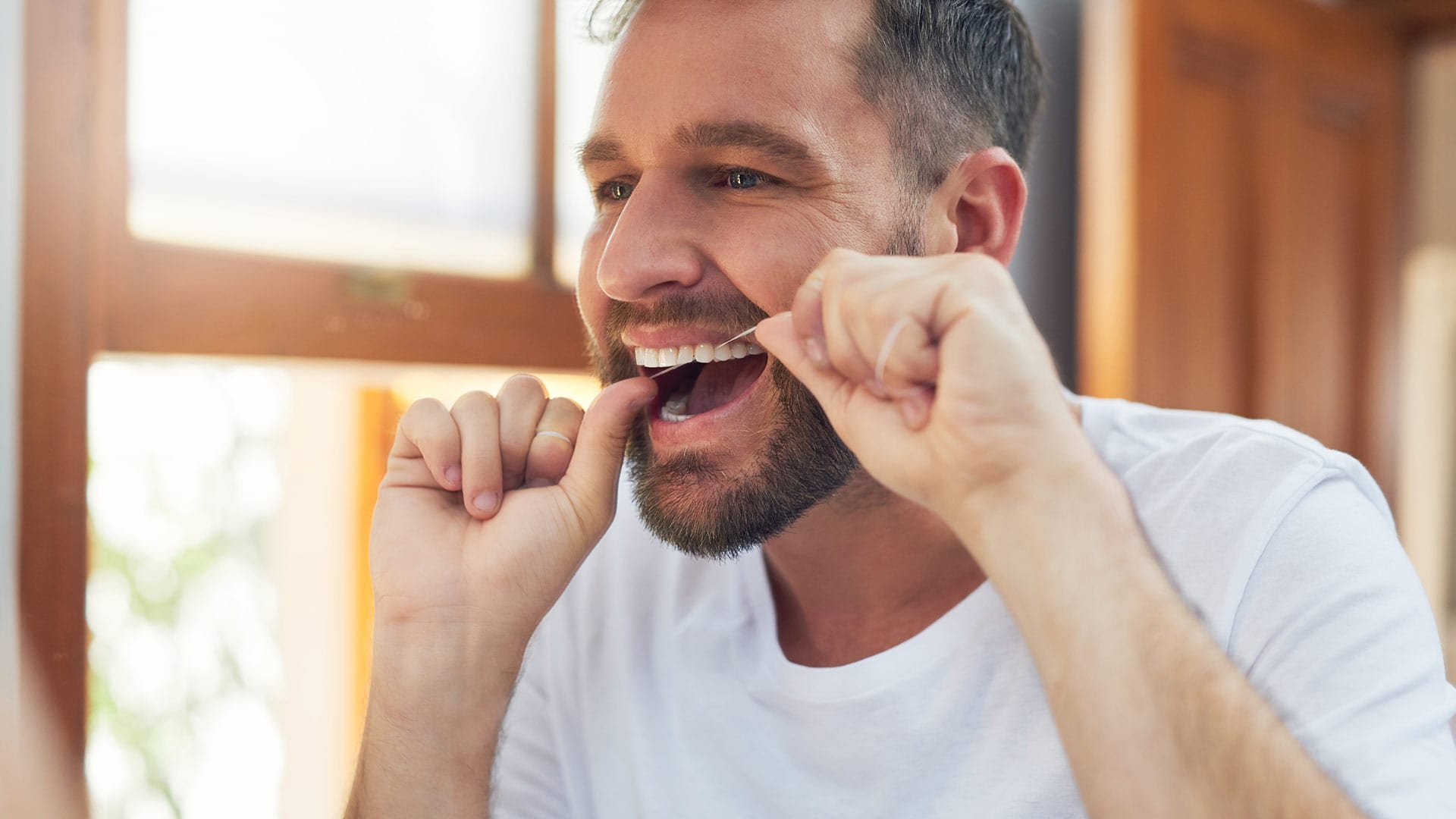 A man with healthy looking teeth and gums practices good dental hygiene by flossing in front of a mirror.